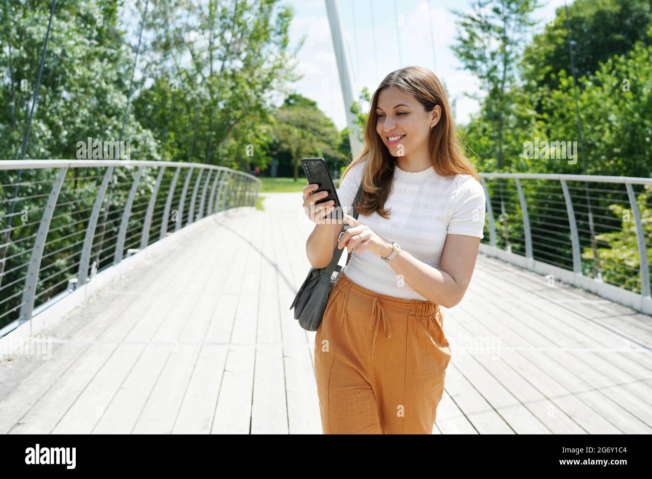 Glückliche junge Frau mit bequemen Kleidern, die mit dem Finger auf dem Smartphone im Stadtpark tippen. Nachhaltige umweltfreundliche Stadt- und Lifestyle-Technologie Konz Stockfoto