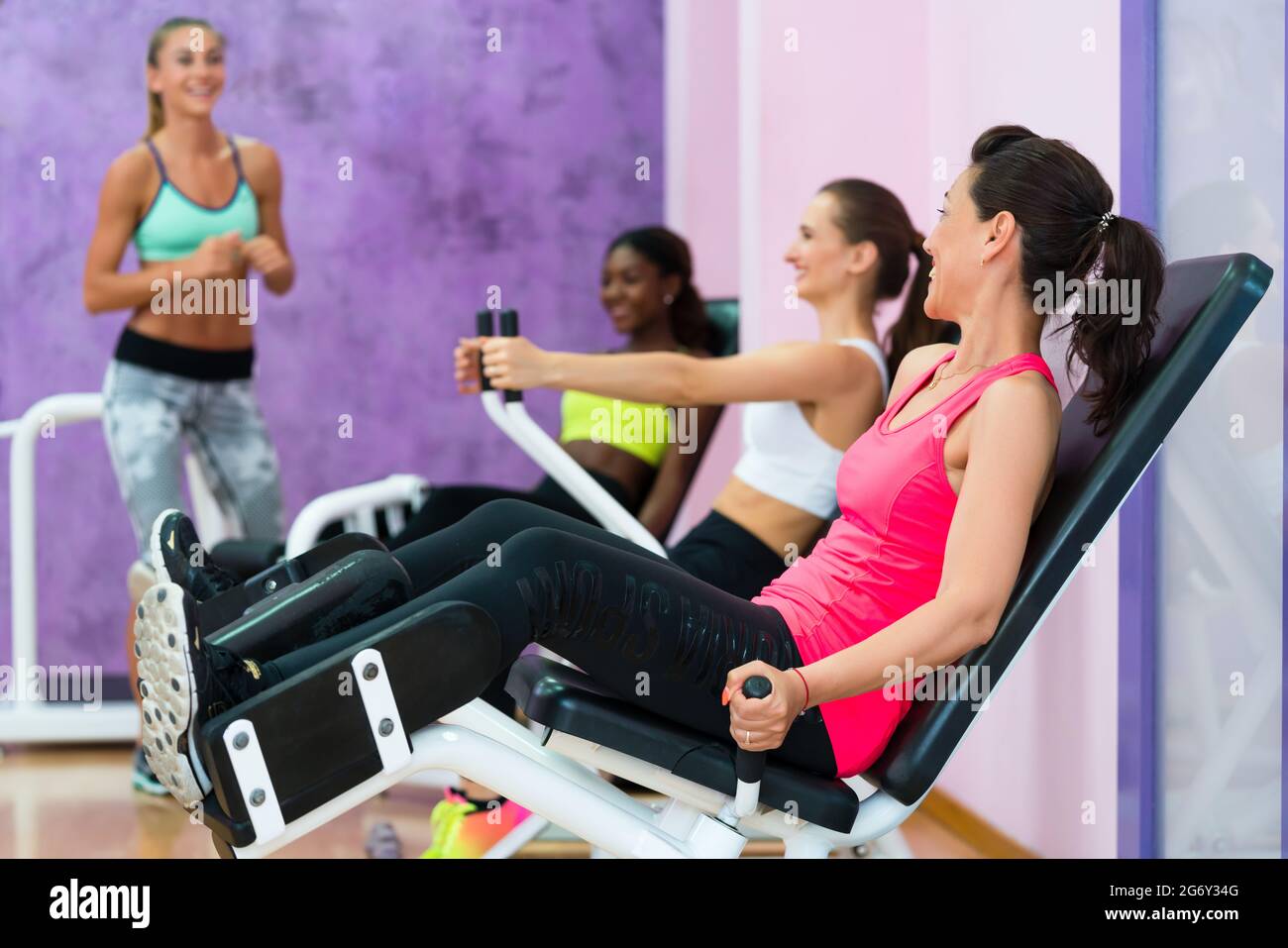 Volle Seitenansicht einer fittenden Frau, die lacht, während sie den Anweisungen eines qualifizierten Fitnesstrainers lauscht, mit Sinn für Humor in der Gruppe Stockfoto