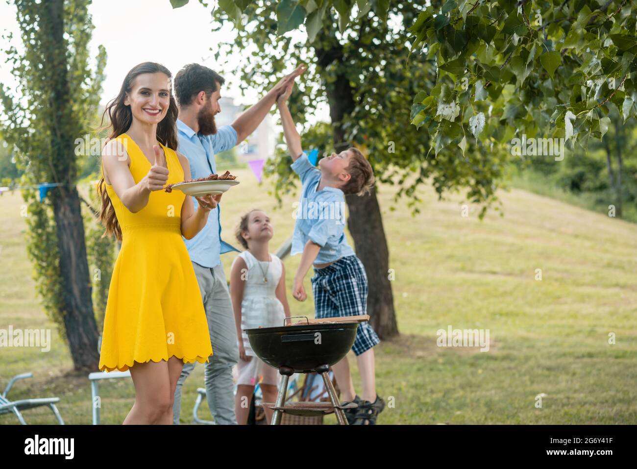 Wunderschöne Frau, Mutter von zwei Kindern, lächelnd und mit Daumen nach oben, während sie im Sommer mit ihrer glücklichen Familie beim BBQ verbringt Stockfoto