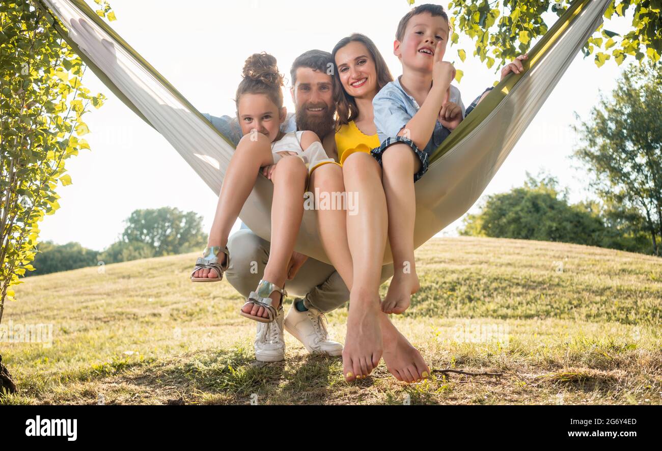 Family Portrait mit einer schönen Mutter von zwei Verspielte Kinder schwingen in der Hängematte, während bei camera Suchen neben ihrem Ehemann im Freien im Sommer Stockfoto