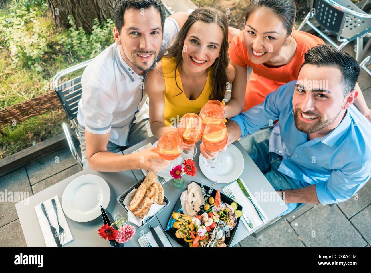 Ein Porträt aus dem Blickwinkel von vier fröhlichen und schönen jungen Freunden, die in einem trendigen Restaurant mit köstlichen Speisen in die Kamera schauen und dabei die Kamera anstoßen Stockfoto