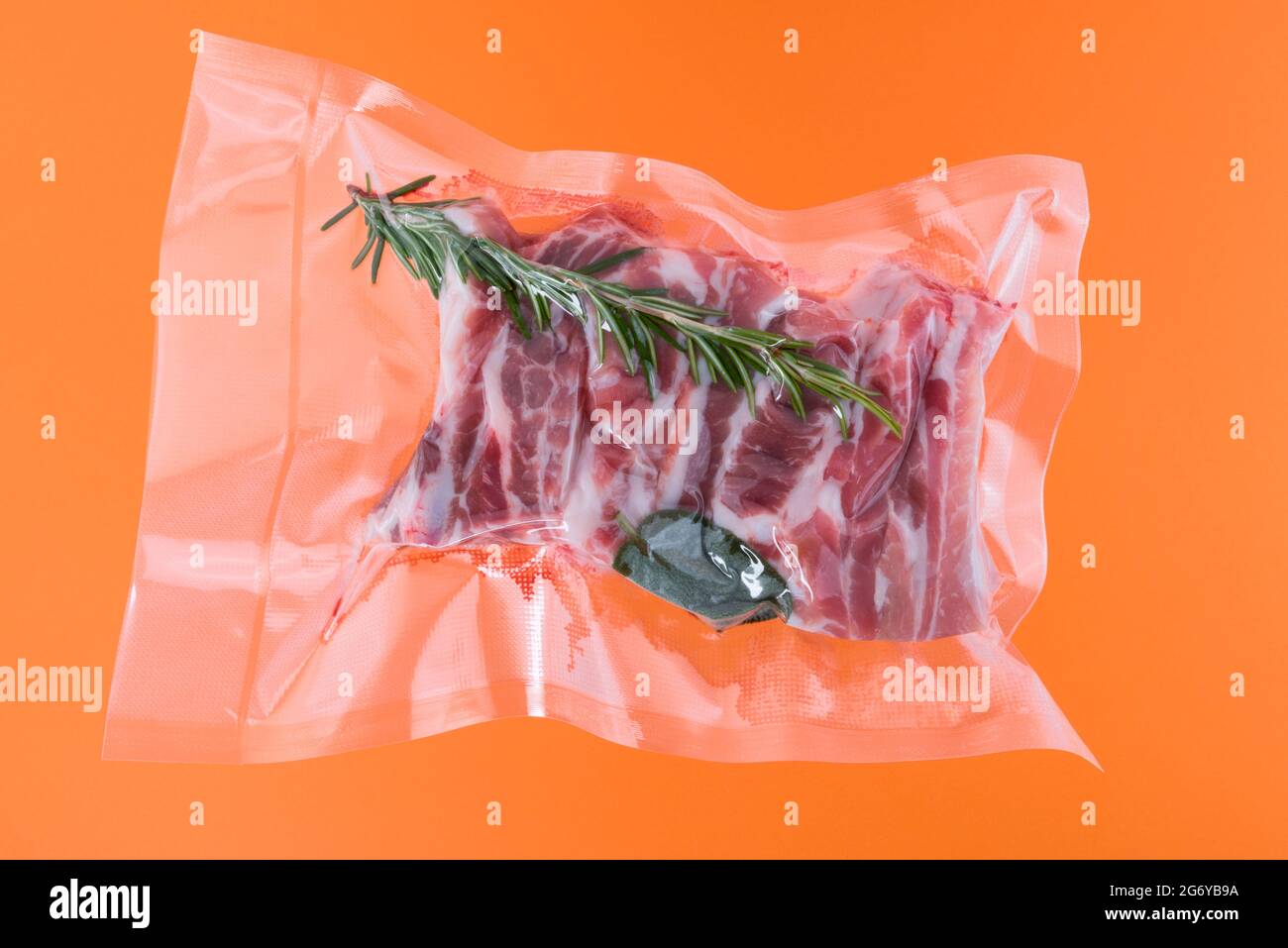 Schweineribs mit Rosmarin in vakuumverpackt, versiegelt für Sous-Vide-Kochen, isoliert auf orangefarbenem Hintergrund Stockfoto