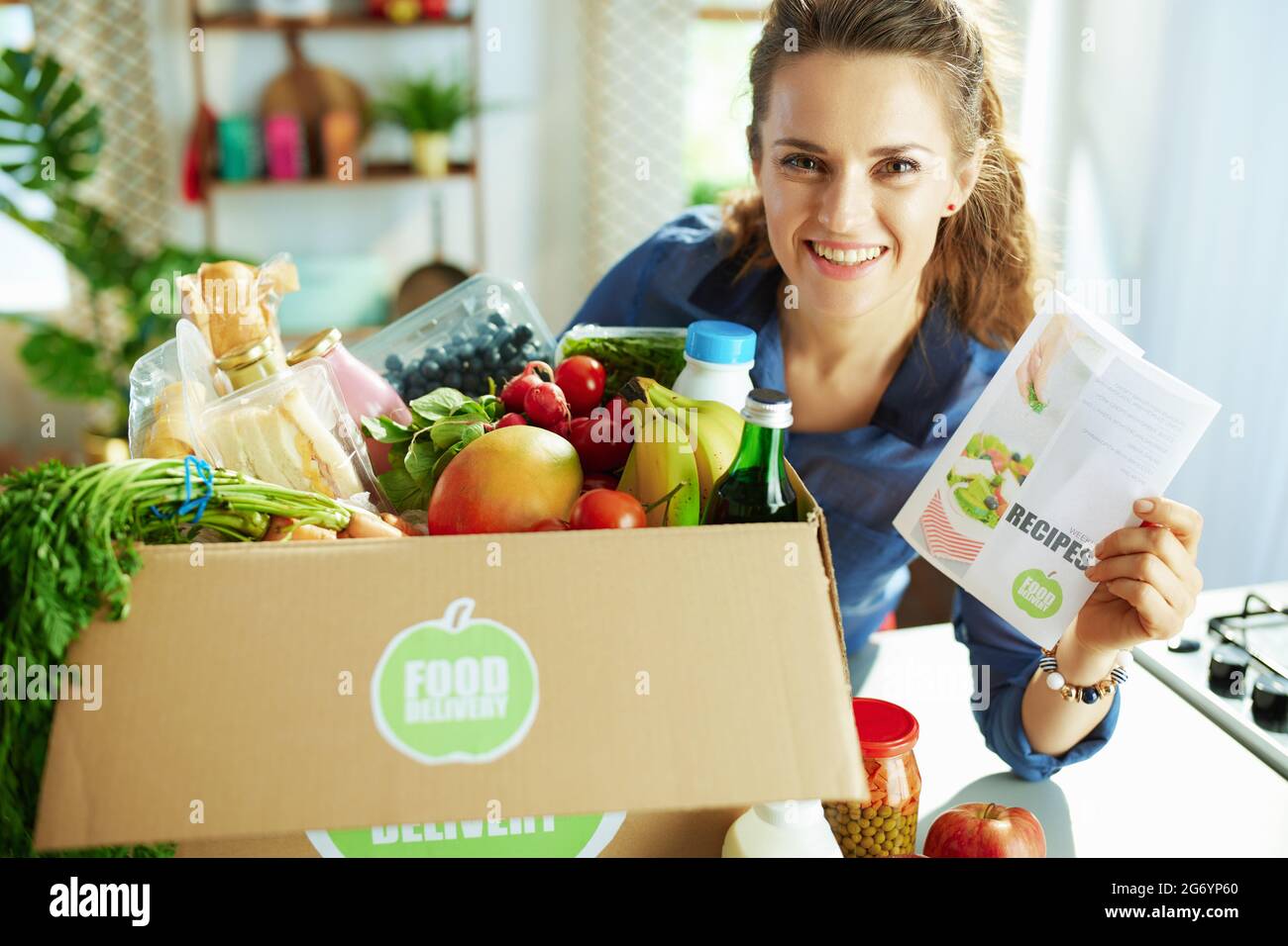 Lieferung von Lebensmitteln. Glückliche moderne Frau mit Lebensmittel-Box und Rezepte-Liste in der Küche. Stockfoto