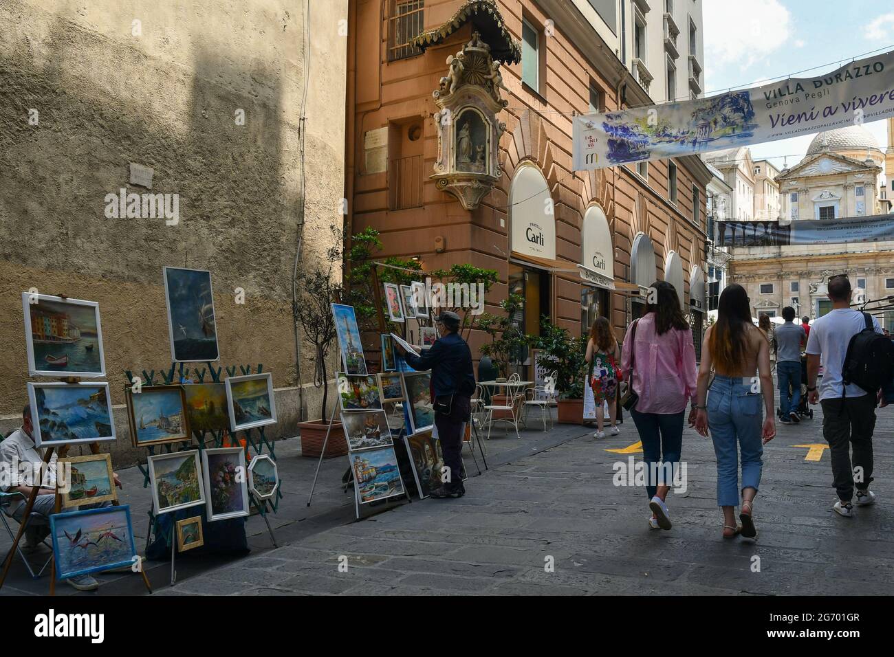 Straßenkünstler verkaufen ihre Gemälde in der Via San Lorenzo im historischen Zentrum von Genua mit Touristen, die im Sommer, Ligurien, Italien, spazieren Stockfoto