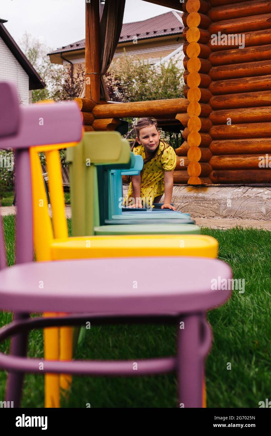 Ein fröhliches Mädchen kriecht entlang einer Reihe von bunten Holzstühlen, ein Mädchen hat eine gute Zeit im Hinterhof Stockfoto