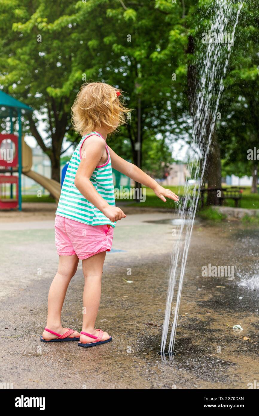 Ein kleines, glückliches Kind, das am heißen Sommertag auf dem Splash Pad im örtlichen öffentlichen Park mit Wasser spielt. Kleine schöne Mädchen mit Spaß am Brunnen Spielplatz. Stockfoto