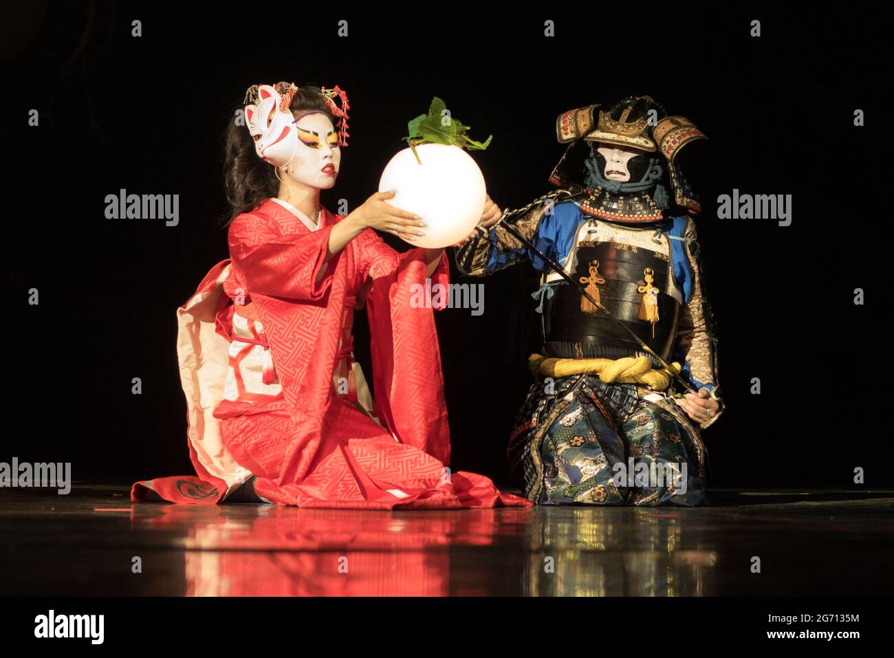 Japanische Geisha in traditionellem Kimono und Fuchsmaske mit Kugellampe und Samurai-Krieger in Rüstung sitzen im Dunkeln auf den Knien. Traditionell Stockfoto