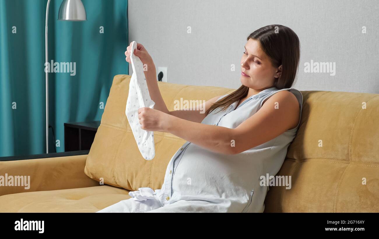 Eine Schwangere mit großem Bauch bereitet Babykleidung vor, während sie auf dem Sofa sitzt. Stockfoto