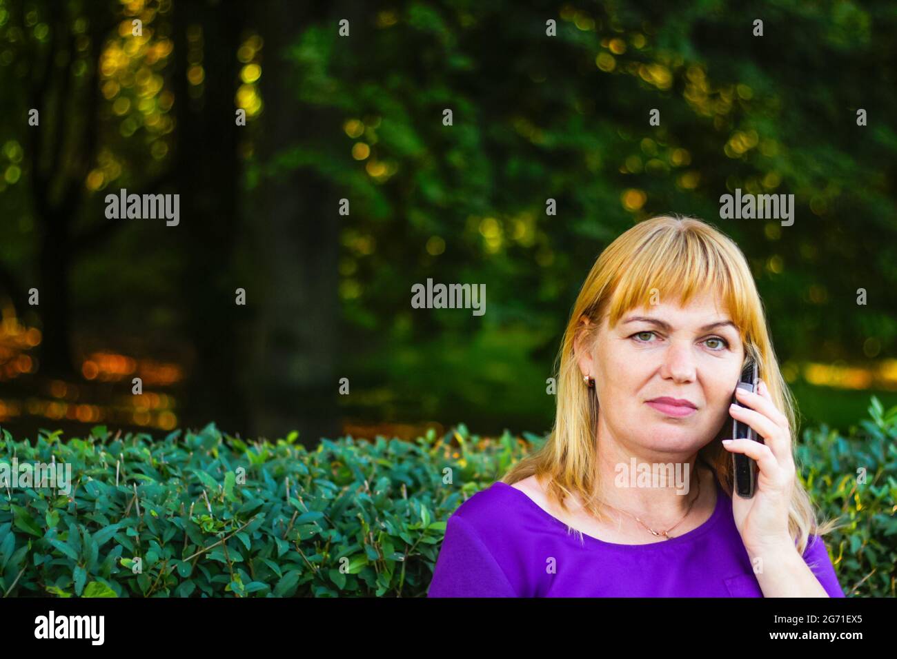 Unschärfe kaukasisch blonde Frau reden, sprechen am Telefon draußen, im Freien. 40-jährige Frau in lila Bluse im Park auf der Bank. Erwachsene, weiblich pro Stockfoto