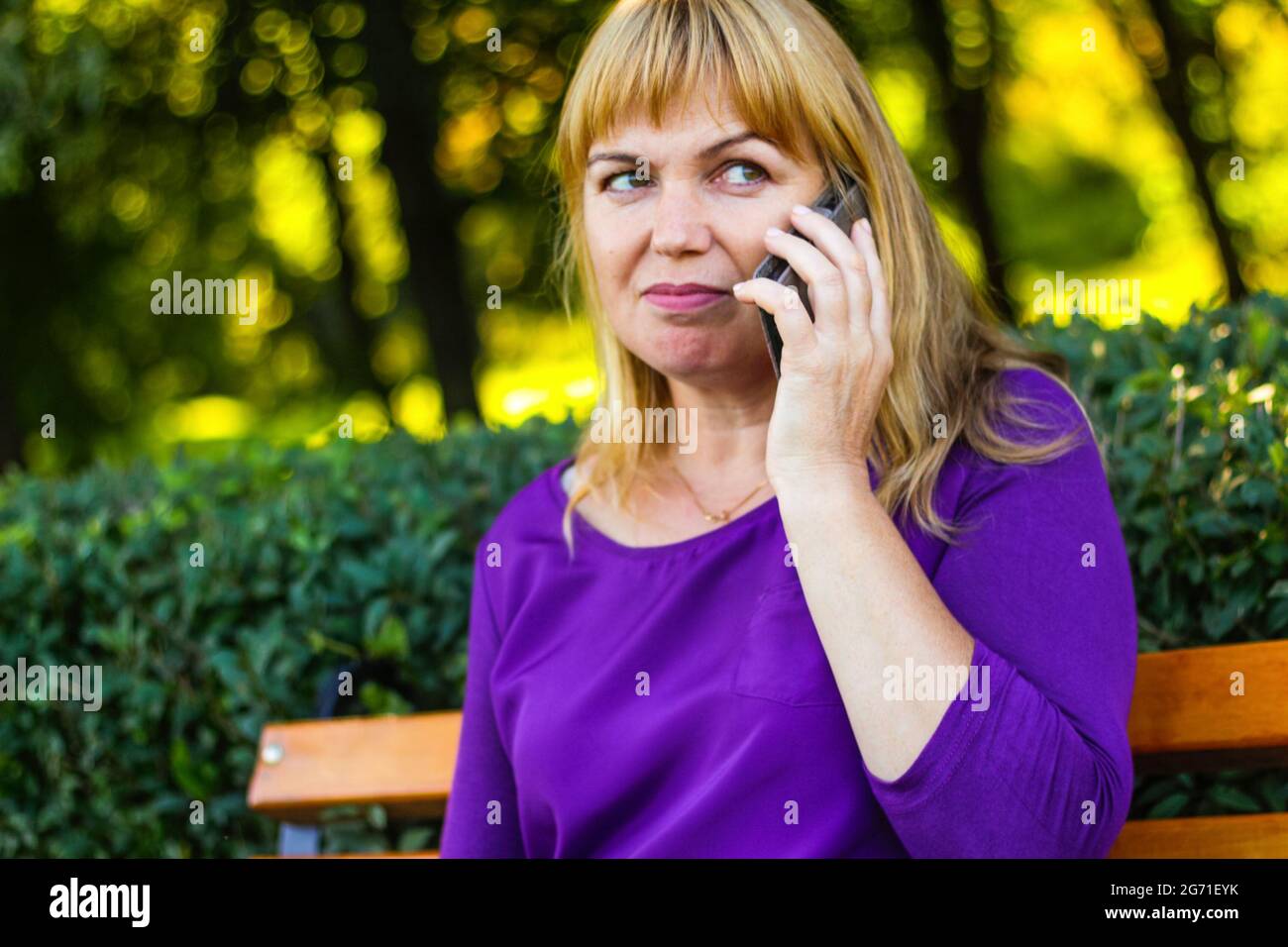 Unschärfe kaukasisch blonde Frau sprechen auf dem Telefon draußen, draußen. 40-jährige Frau in lila Bluse im Park auf der Bank. Nette Person mit Handy, Stockfoto