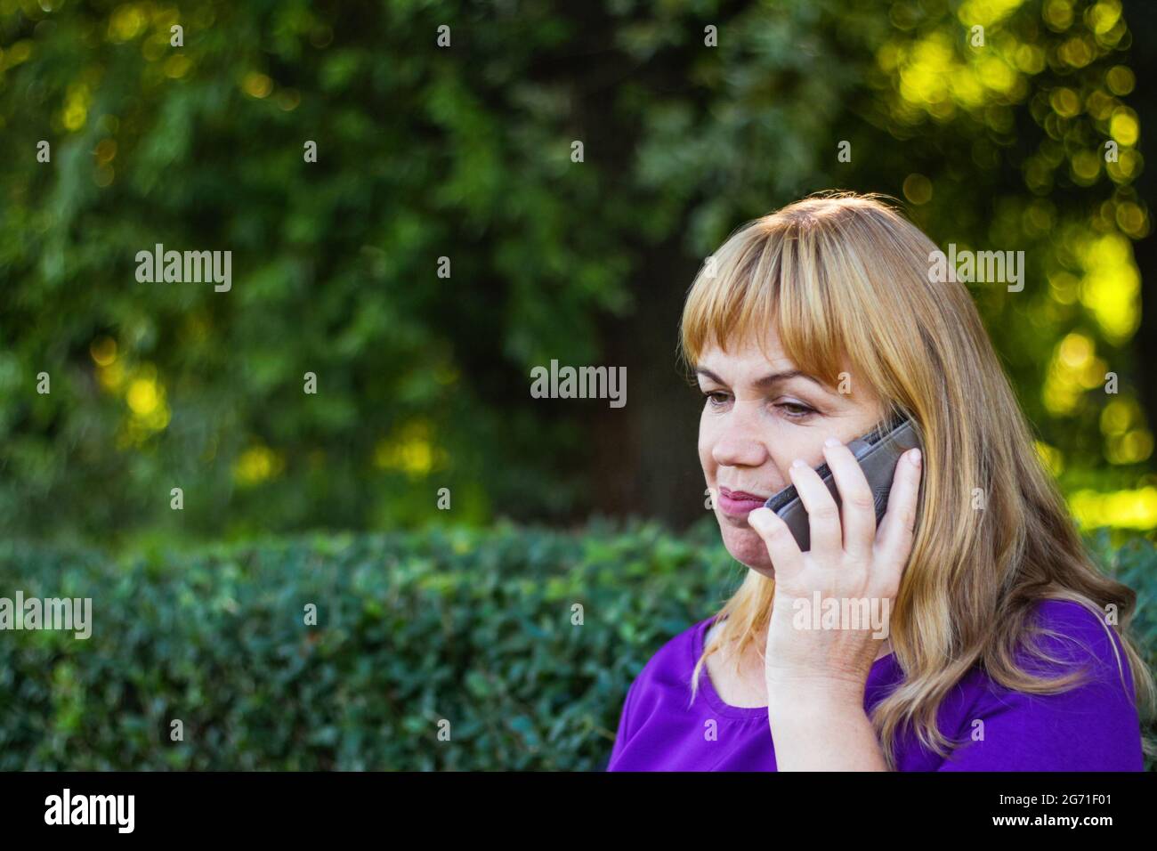 Unschärfe kaukasisch blonde Frau sprechen auf dem Telefon draußen, draußen. 40-jährige Frau in lila Bluse im Park. Seriöse Person mit mobilen, smartp Stockfoto