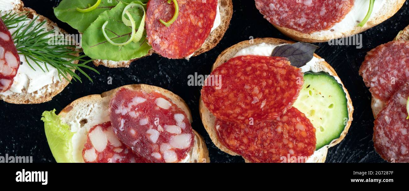 Banner mit Nahlandbrötchen oder Snacks mit verschiedenen Würstchen und Gemüse auf schwarzem Hintergrund. Weicher Fokus. Draufsicht. Flach liegend Stockfoto
