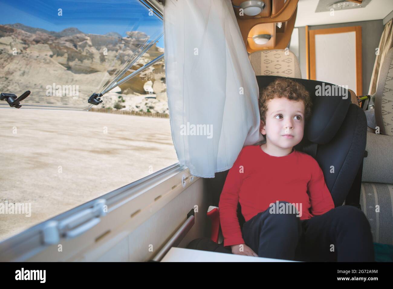 Ein kleiner, lockiger, kaukasisch aussehender Junge, der am offenen Fenster seines geparkten Wohnmobils mit einer spektakulären Aussicht sitzt. Stockfoto