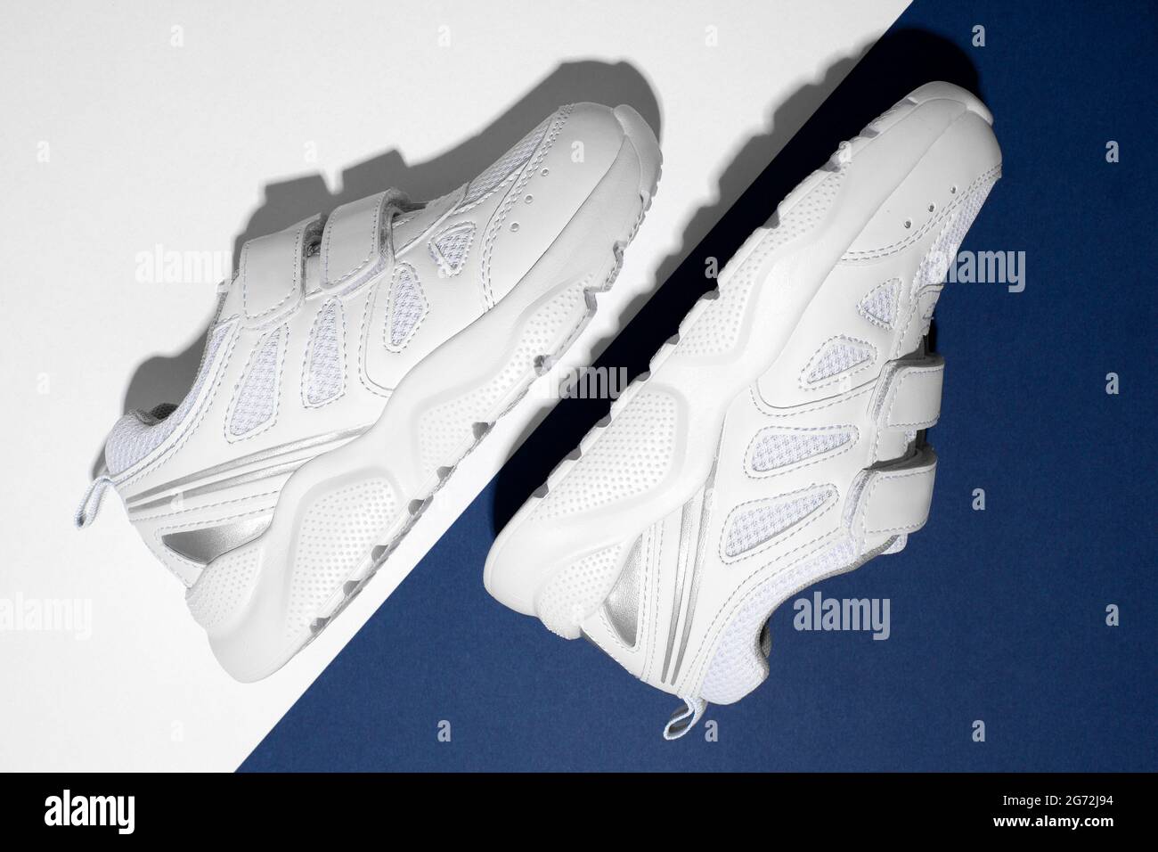 Draufsicht MACRO zwei weiße Kinder-Sneakers an der Seite mit Klettverschlüssen für bequemes Schuhwerk auf einem modernen blau-weißen Papierhintergrund mit Stockfoto
