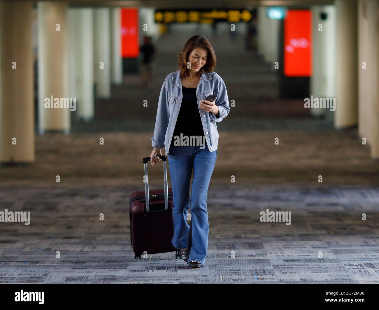 Schöner asiatischer Reisender, der in legerer Kleidung mit Smartphone und zu Fuß im Flughafenterminal unterwegs ist. Stockfoto