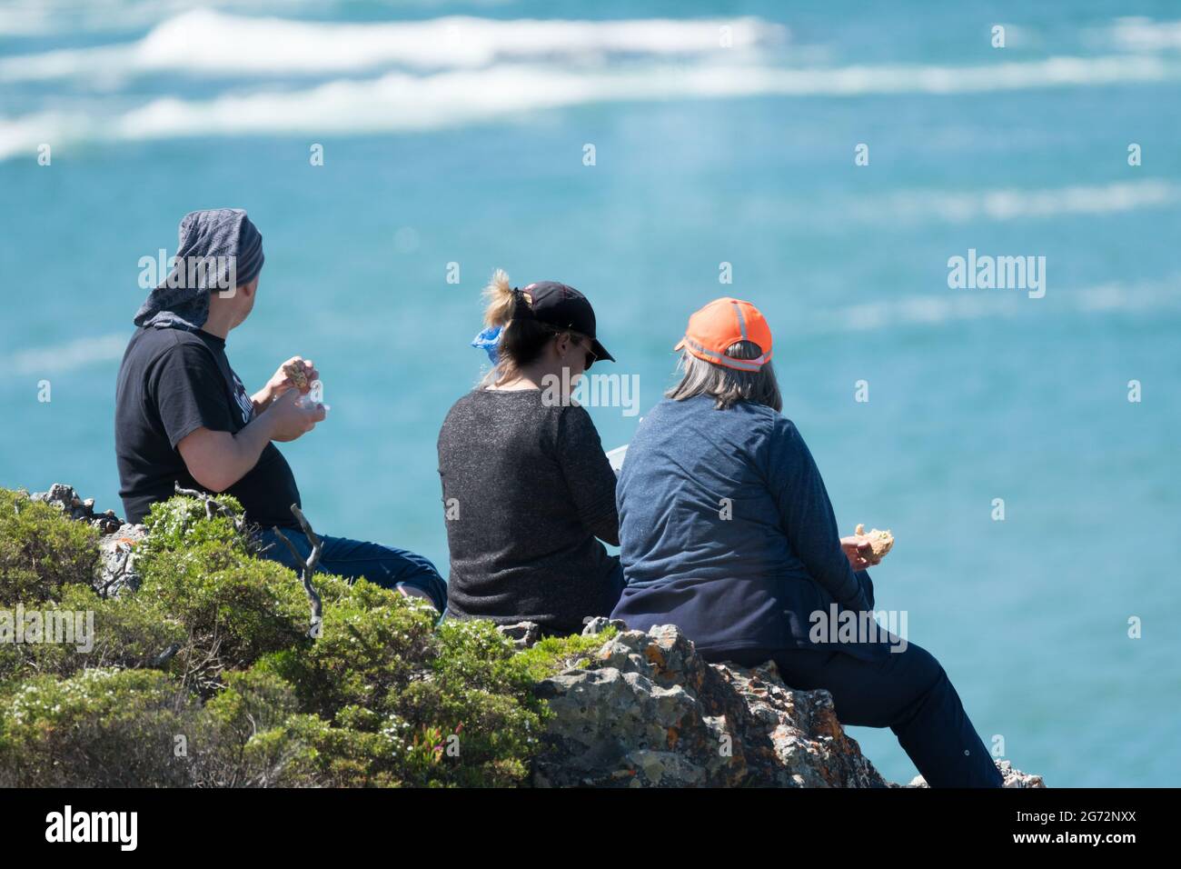 Eine Gruppe von Menschen, die auf den Felsen sitzen und das Meer überblicken, essen ein Picknick-Lunch-Konzept Zweisamkeit und Entspannung Stockfoto