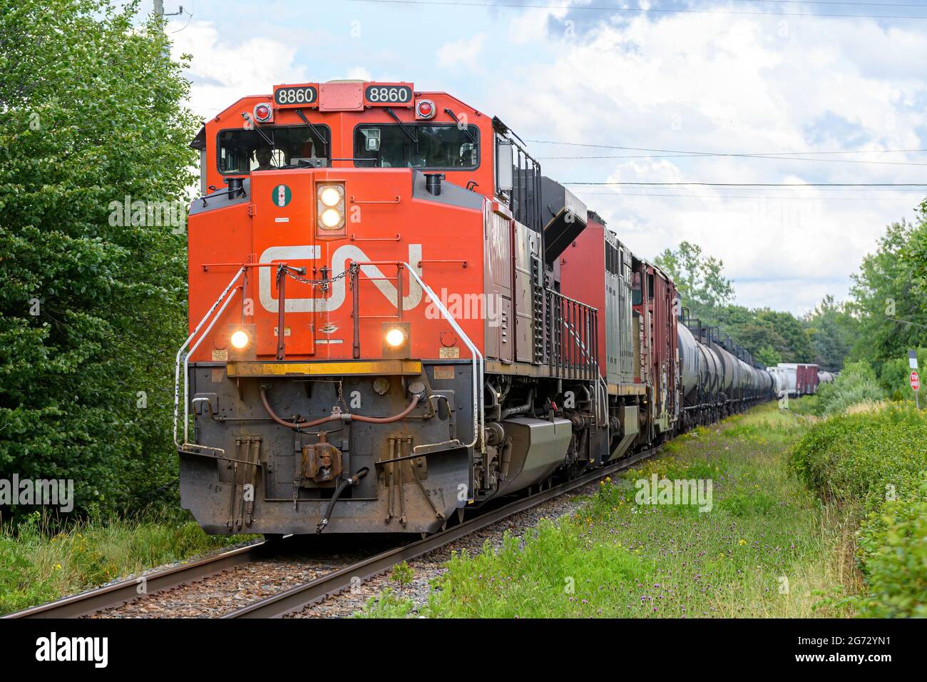 Saint John, NB, Kanada - 6. August 2021: Ein Zug der Canadian National Railway auf einem bewölkten Sagen. Lichter sind an, Bäume und Gras um die Schienen herum. Stockfoto