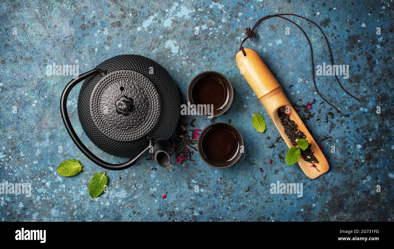 Draufsicht auf schwarze japanische gusseiserne Teekannen und Tee in Bambuslöffel mit grünen Minzblättern auf blauem Beton Hintergrund Stockfoto