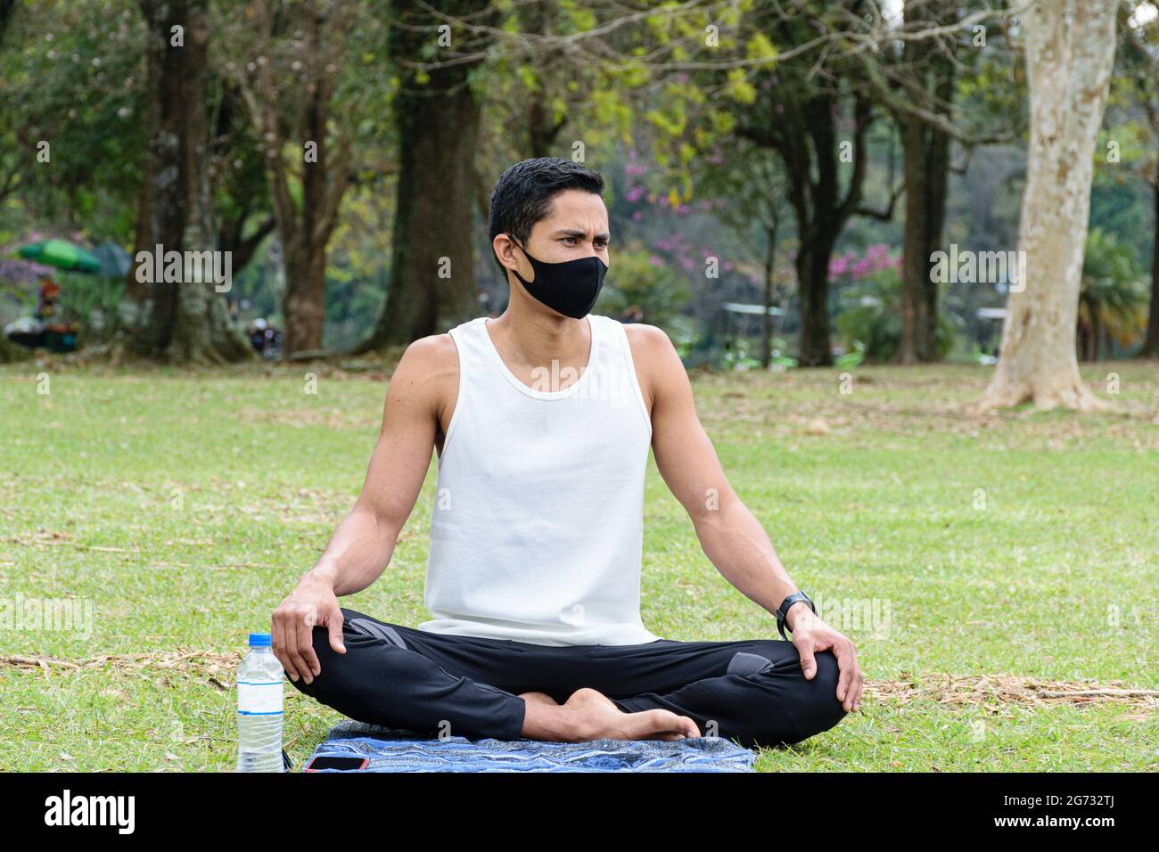 Der 28-jährige brasilianische junge Mann, Yogalehrer, sitzt auf dem Rasen neben einer Flasche Wasser (Vorderansicht). Stockfoto