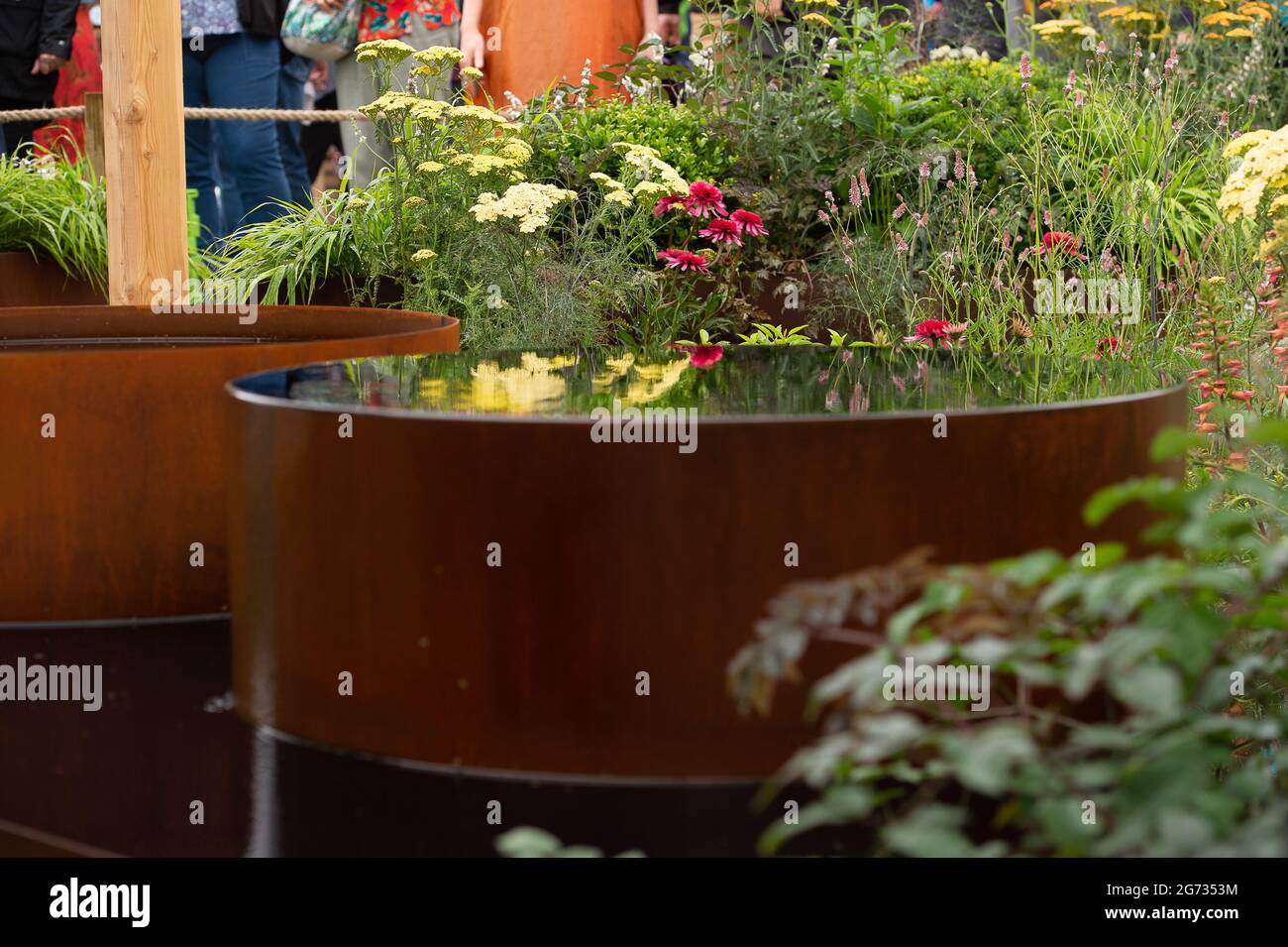 East Molesey, Surrey, Großbritannien. Juli 2021. Einer der Global Impact Gardens - der Fashion Footprint Garden. Designer Baz Grainger sprach heute mit Gärtnern über sein Design. Der Kies im Garten besteht aus weggeworfenen Knöpfen. Die beiden Fässer aus Cortenstahl erinnern an die Farbstoffbehälter, die beim Indigofärben verwendet werden. Quelle: Maureen McLean/Alamy Stockfoto