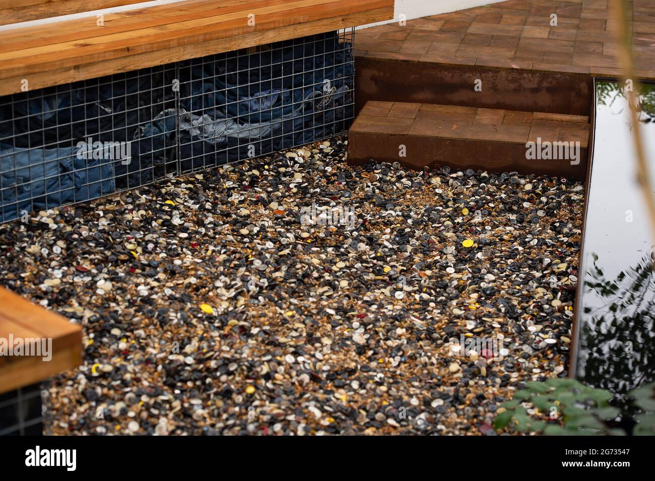 East Molesey, Surrey, Großbritannien. Juli 2021. Einer der Global Impact Gardens - der Fashion Footprint Garden. Designer Baz Grainger sprach heute mit Gärtnern über sein Design. Der Kies im Garten besteht aus weggeworfenen Knöpfen. Die beiden Fässer aus Cortenstahl erinnern an die Farbstoffbehälter, die beim Indigofärben verwendet werden. Quelle: Maureen McLean/Alamy Stockfoto