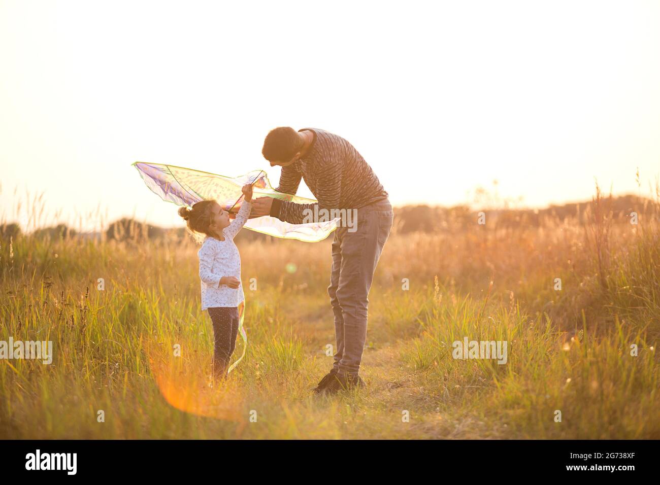 Papa hilft seiner Tochter, im Sommer bei Sonnenuntergang einen Drachen auf einem Feld zu fliegen. Familienunterhaltung im Freien, Vatertag, Kindertag. Ländliche Gebiete, zusatzbereich Stockfoto