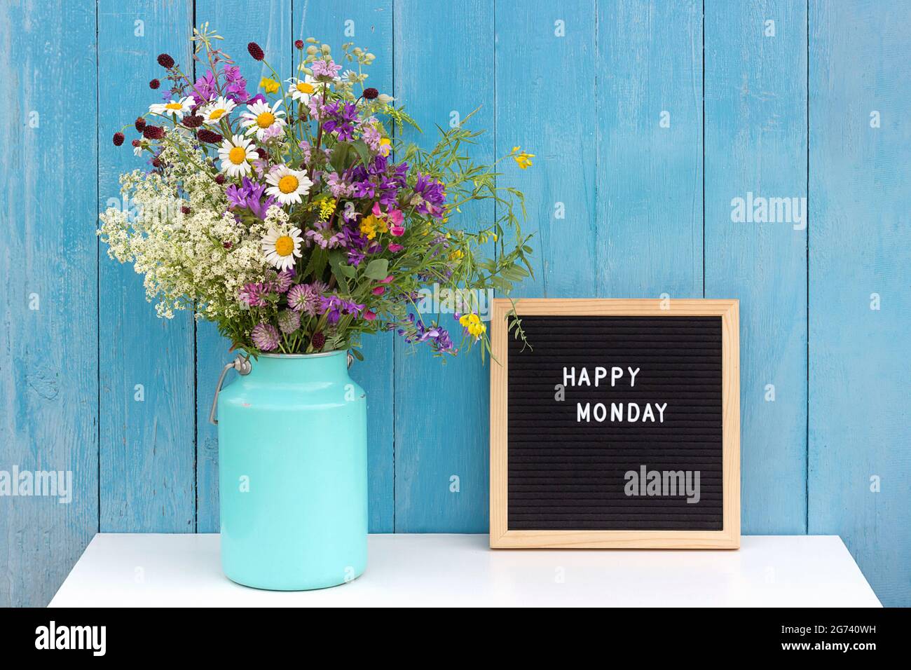 Happy Monday Worte auf schwarzem Briefbrett und Bouquet von hellen wilden Blumen in Zinn können Vase auf Tisch gegen blaue Holzwand. Konzept Hallo Montag. Stockfoto