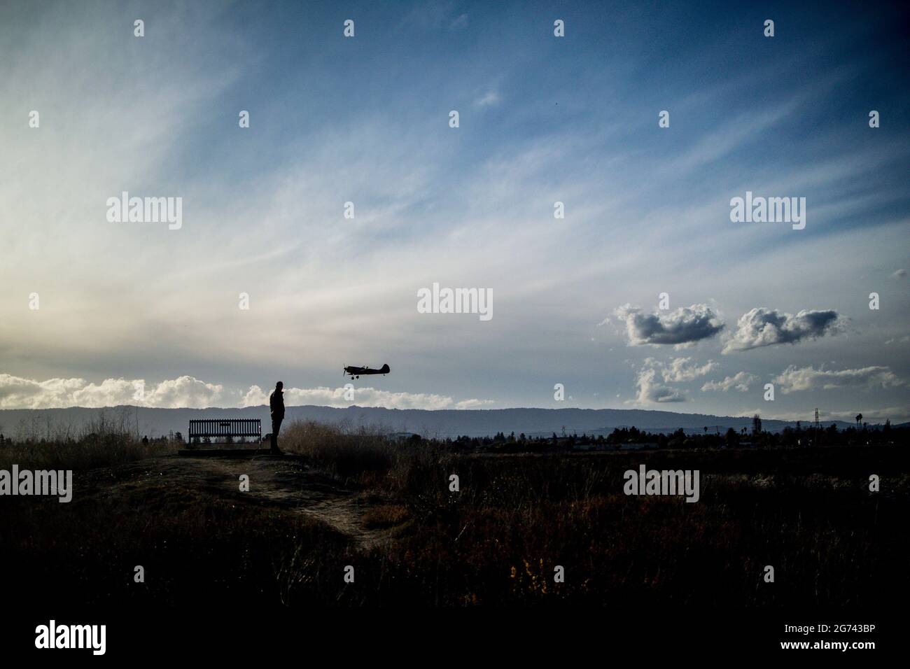 Silhouette eines Mannes, der neben einer Bank steht, mit einem kleinen Flugzeug, das tief über seinem Kopf fliegt, Berge am Horizont, blauer Himmel mit Wolken Stockfoto