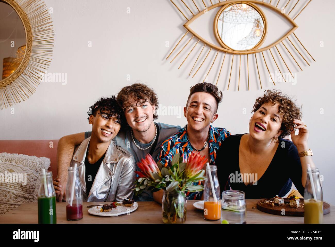Kreis glücklicher Freunde, die in einem Restaurant zusammen sitzen. Vier junge, schwule Menschen lächeln beim Mittagessen fröhlich vor der Kamera. Freunde verbinden und s Stockfoto