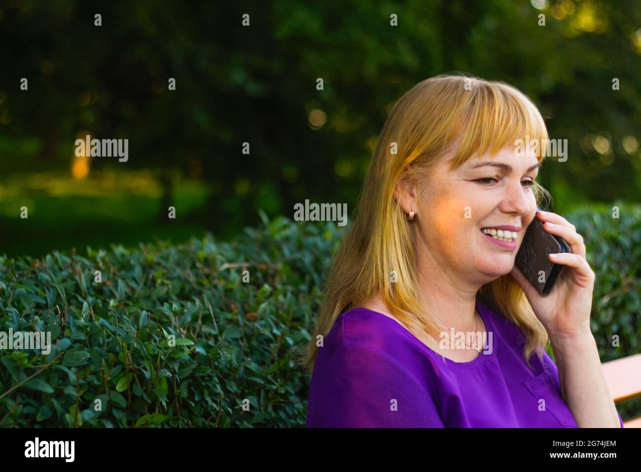 Unschärfe-Nahaufnahme lächelnde kaukasische blonde Frau, die im Freien telefoniert. 40-jährige Frau in lila Bluse im Park. Erwachsene, pe Stockfoto