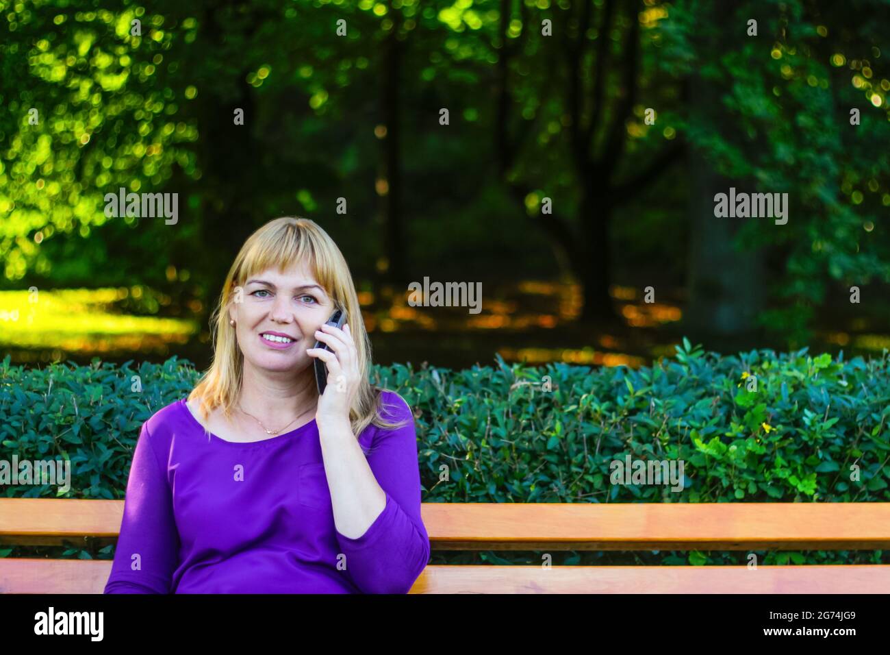 Unschärfe kaukasisch blonde lächelnde Frau sprechen, sprechen auf dem Telefon draußen, draußen. 40-jährige Frau in lila Bluse im Park auf der Bank. Erwachsene fe Stockfoto