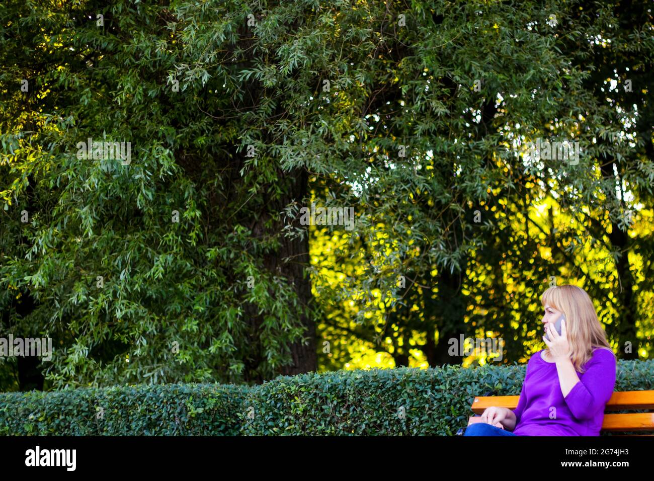 Unschärfe kaukasisch blonde Frau reden, sprechen am Telefon draußen, im Freien. 40-jährige Frau in lila Bluse im Park. Erwachsene Frauen, die ein Mobiltelefon verwenden Stockfoto
