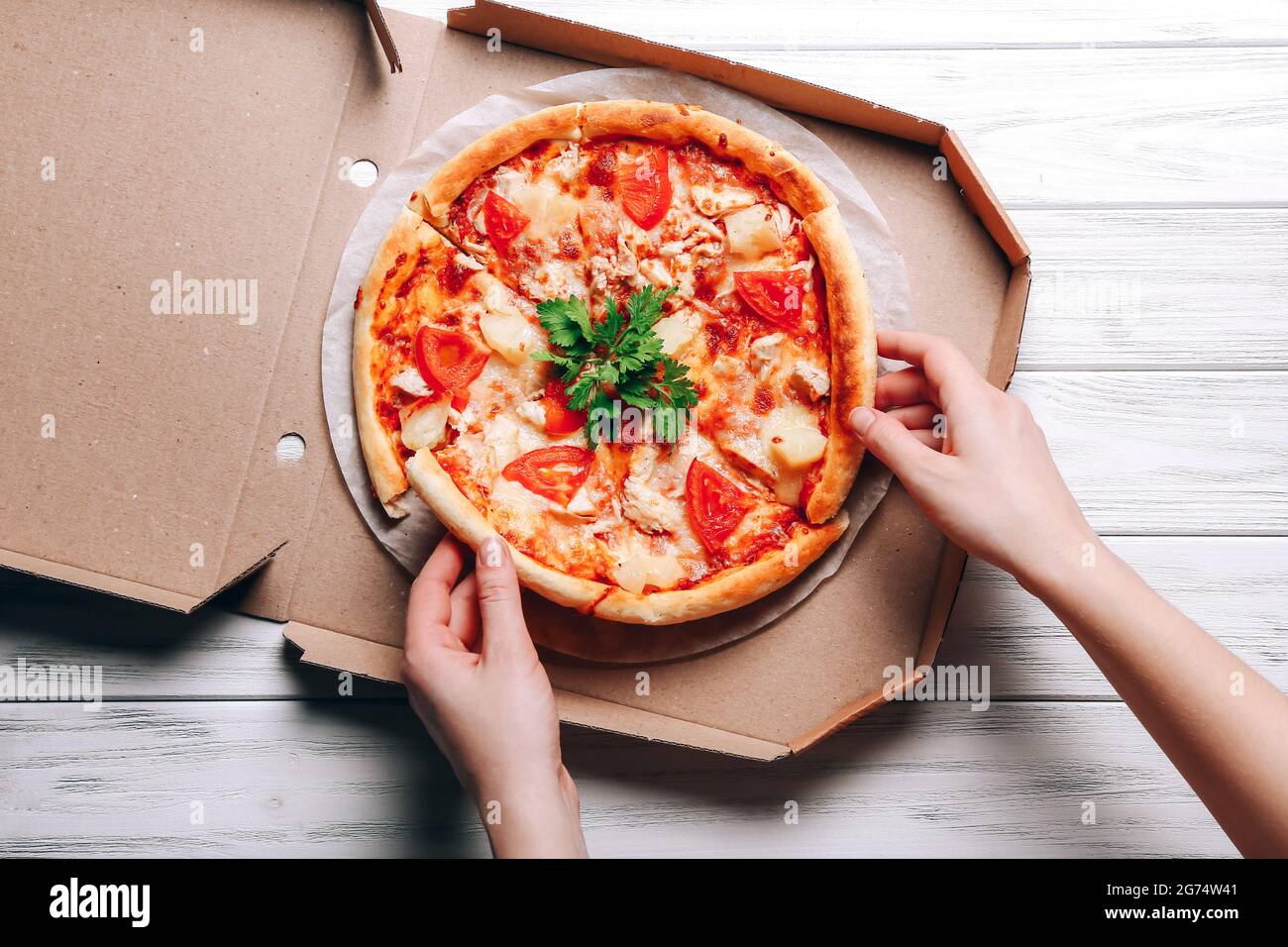 Leckere Pizza im Lieferkarton und Frau Hände nehmen Pizzascheiben auf weißem Holzhintergrund Stockfoto