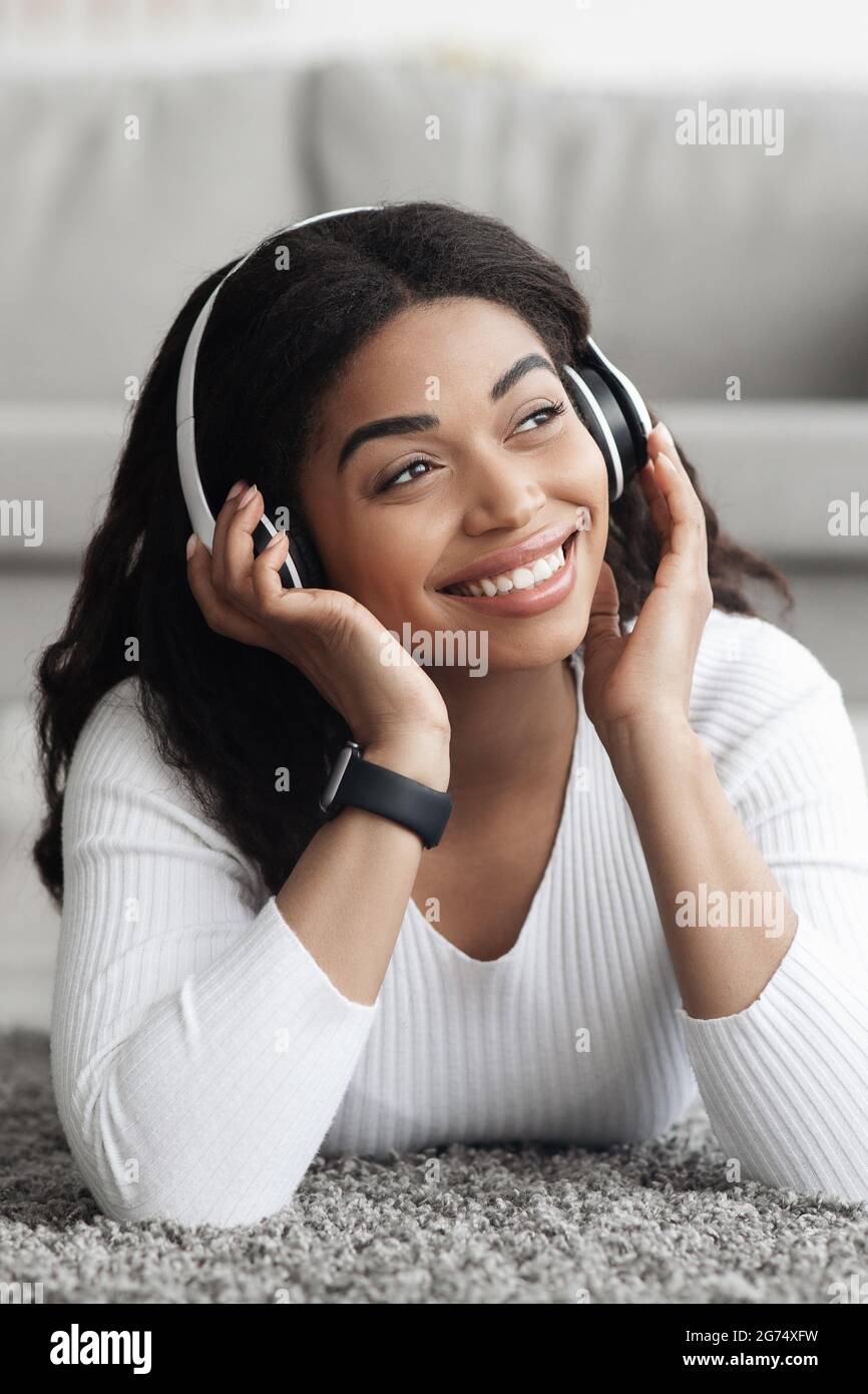 Porträt einer glücklichen schwarzen Frau in kabellosen Kopfhörern, die Lieblingssongs hört, auf dem Bodenteppich liegt, sich ausschneiden lässt Stockfoto