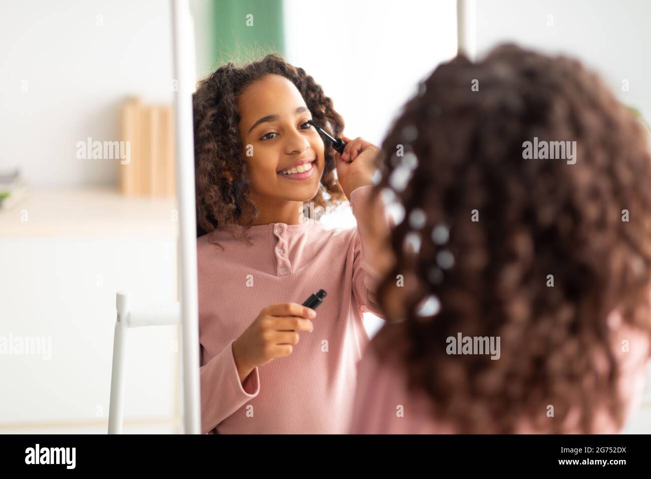 Make-up zu Hause. Über die Schulter des schwarzen Teenagers, das schwarze Mascara auftradet, den Spiegel anschaut und lächelt Stockfoto