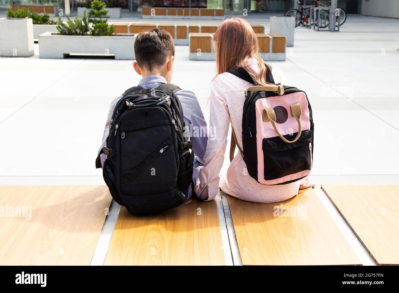 Rückansicht von zwei kaukasischen Schülerinnen und Schülern, die draußen auf einer Holzbank sitzen. Tragen Sie sommerliche Freizeitkleidung, Rucksäcke. Stockfoto