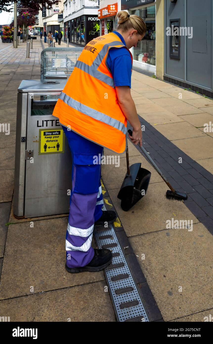 Eine junge Frau, die als Straßenreinigerin Teil des lokalen Pflegeteams der Behörde ist, fegt mit einem kleinen Pinsel eine Fußgängerzone Stockfoto