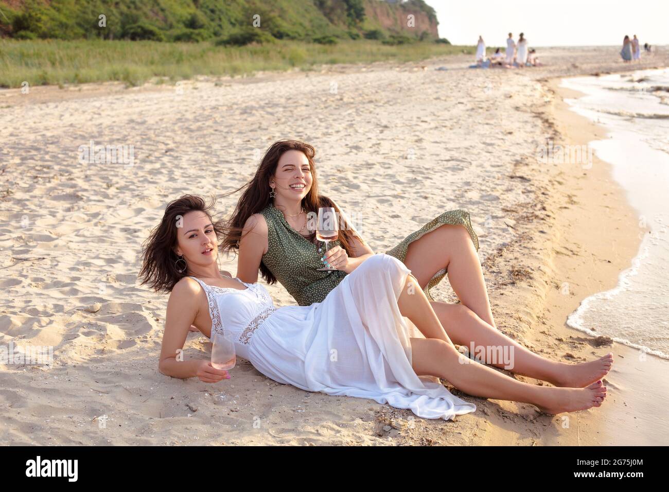 Fröhliche Erwachsene Freundinnen im Sommeroutfit lächeln, während sie Wein am Sandstrand in der Nähe des Meeres genießen Stockfoto