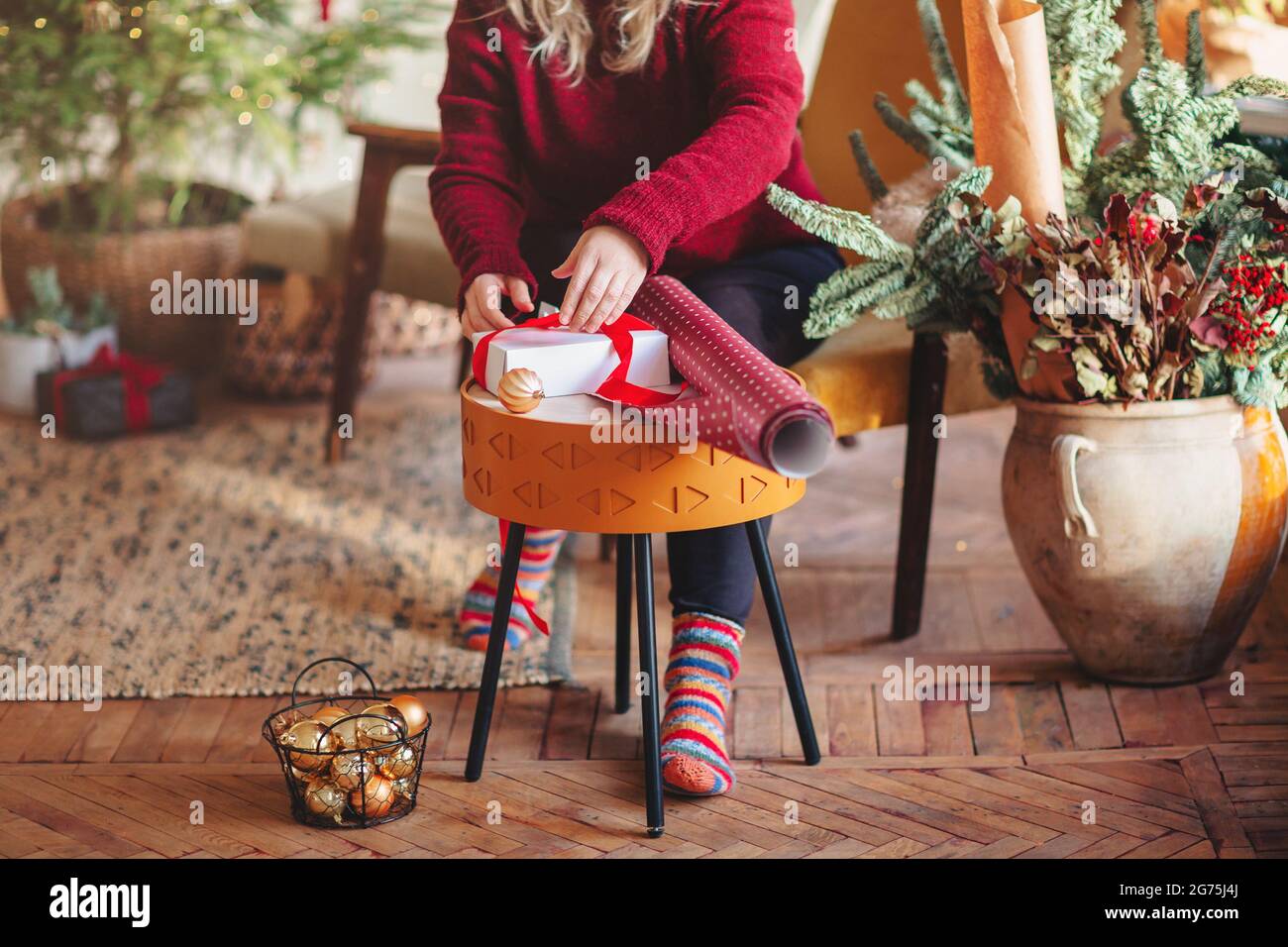 Crop mittleren Alters Frau in lässigem Outfit Verpackung Weihnachtsgeschenk-Box mit Kordel und roten Beeren, während auf Sessel sitzen und mit leichter Girlande dekoriert Stockfoto