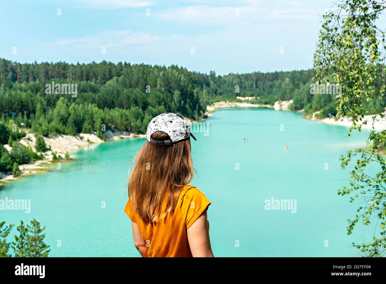 Rückansicht einer jungen blonden Frau, die in einer Mütze und einem Senf-T-Shirt auf der Klippe eines blau-türkisfarbenen Sees oder Flusses und eines Nadelwaldes steht, tr Stockfoto