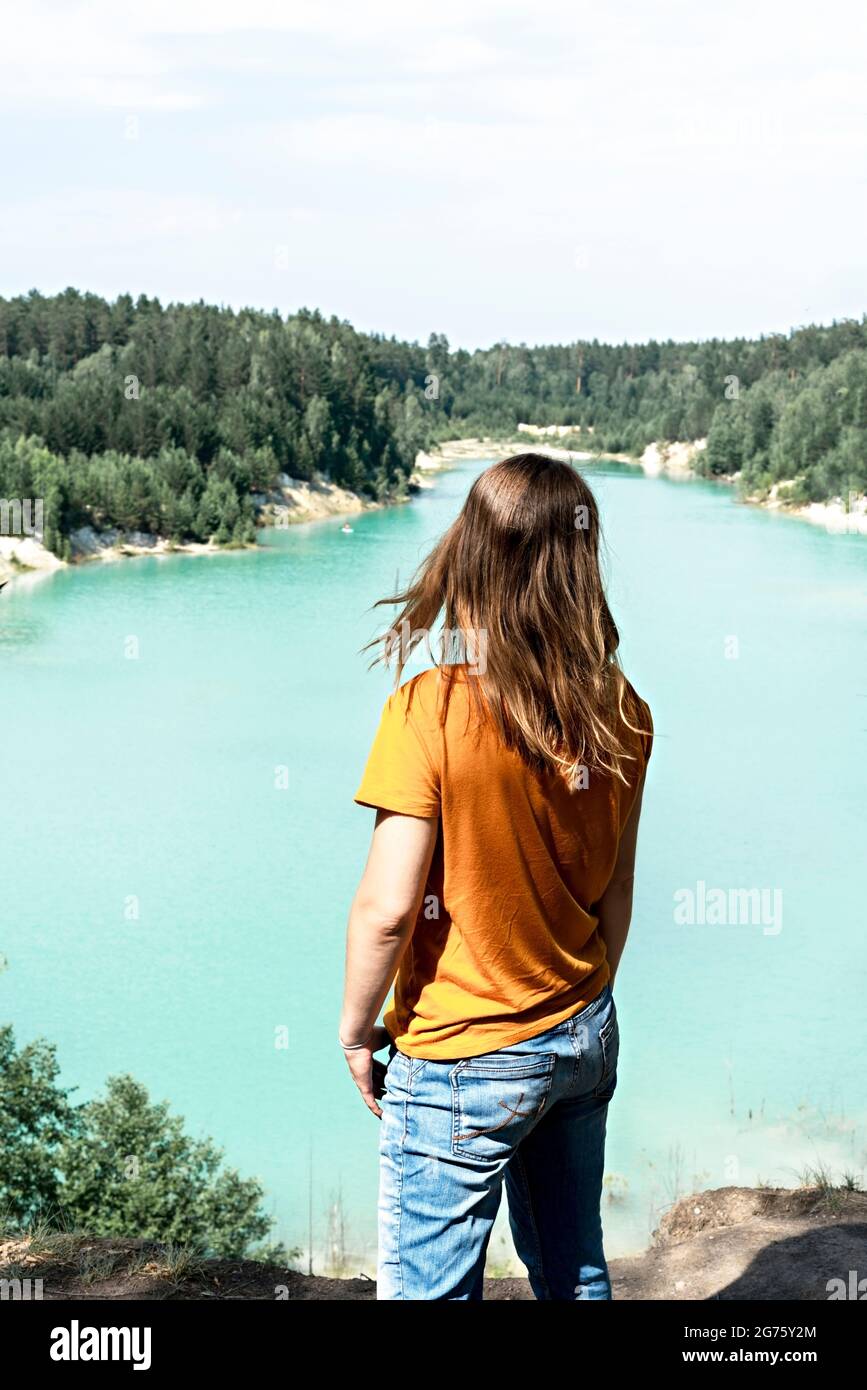 Rückansicht eines jungen blonden Frau Wanderer Senf T-Shirt auf der Klippe des blau türkisfarbenen See oder Fluss und Nadelwald stehen, Reise Natur beau Stockfoto