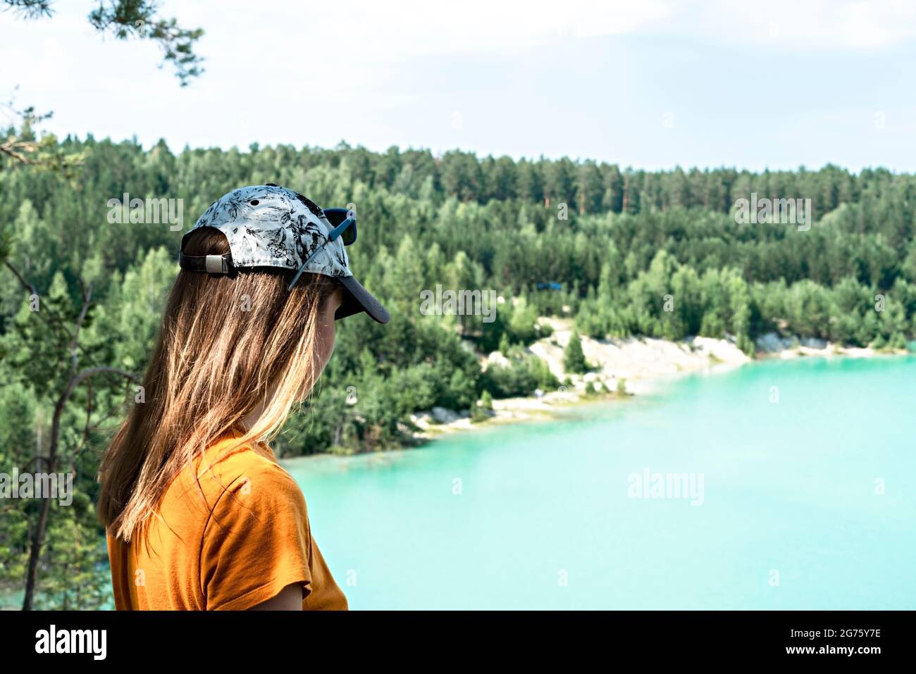 Rückansicht einer jungen blonden Frau, die in einer Mütze und einem Senf-T-Shirt auf der Klippe eines blau-türkisfarbenen Sees oder Flusses und eines Nadelwaldes steht, tr Stockfoto