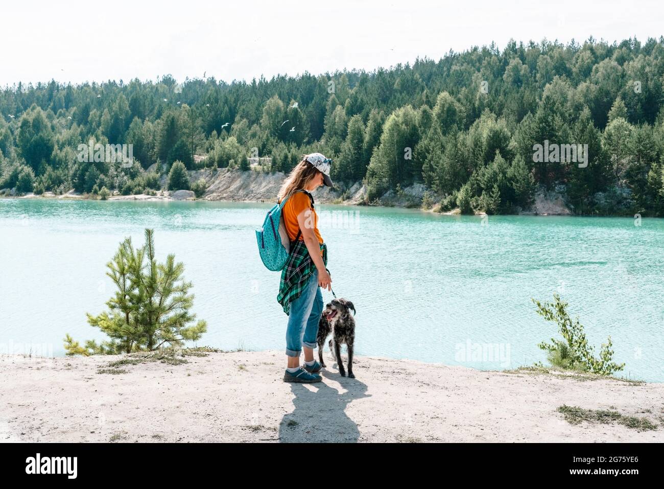 Rückansicht einer jungen Frau, die in einer Mütze mit Rucksack unterwegs ist und am sandigen Ufer des blauen Sees oder des Flusses mit einem flauschigen grauen Hund läuft, Haustier liebt aktiven Lebensstil, t Stockfoto