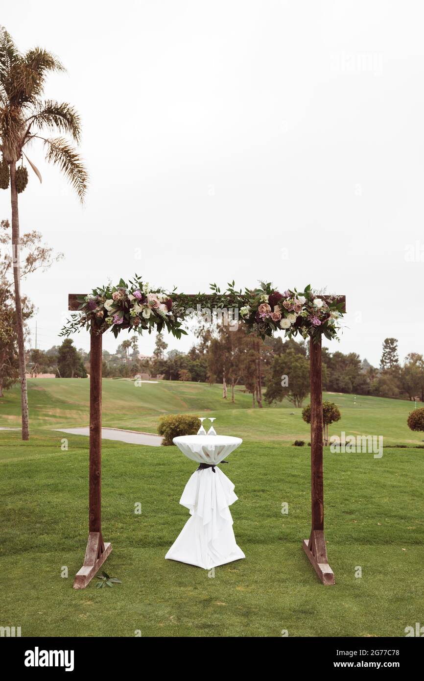 Hochzeitszeremonie im Freien an einem bewölkten Regentag - quadratischer Holzbogen mit Blumenarrangements, weiße Stühle auf grünem Rasen. Stockfoto