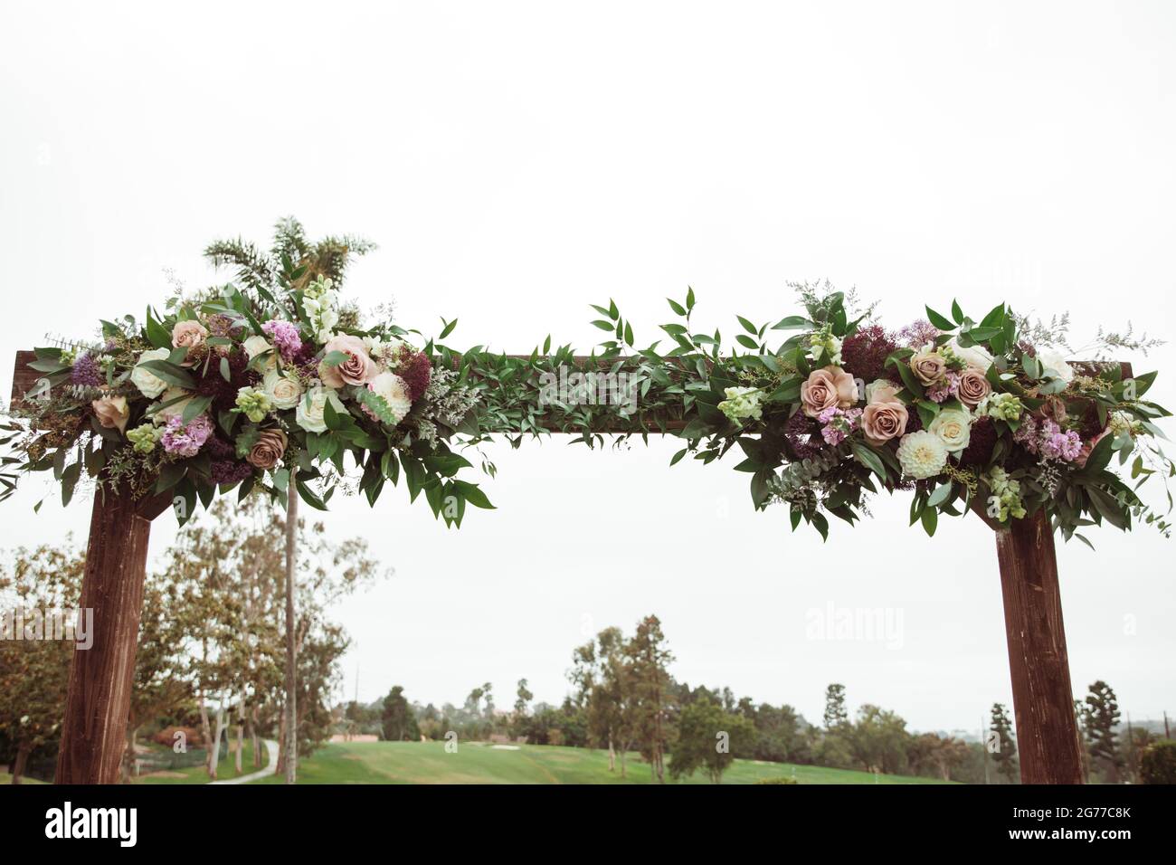 Hochzeitszeremonie im Freien an einem bewölkten Regentag - quadratischer Holzbogen mit Blumenarrangements, weiße Stühle auf grünem Rasen. Stockfoto