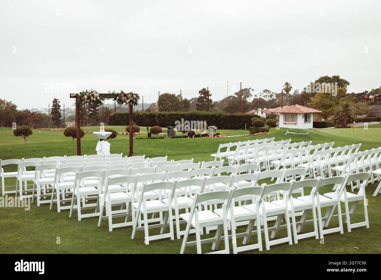 Hochzeitszeremonie im Freien an einem bewölkten Regentag - quadratischer Holzbogen mit Blumenarrangements, weiße Stühle auf grünem Rasen. Stockfoto