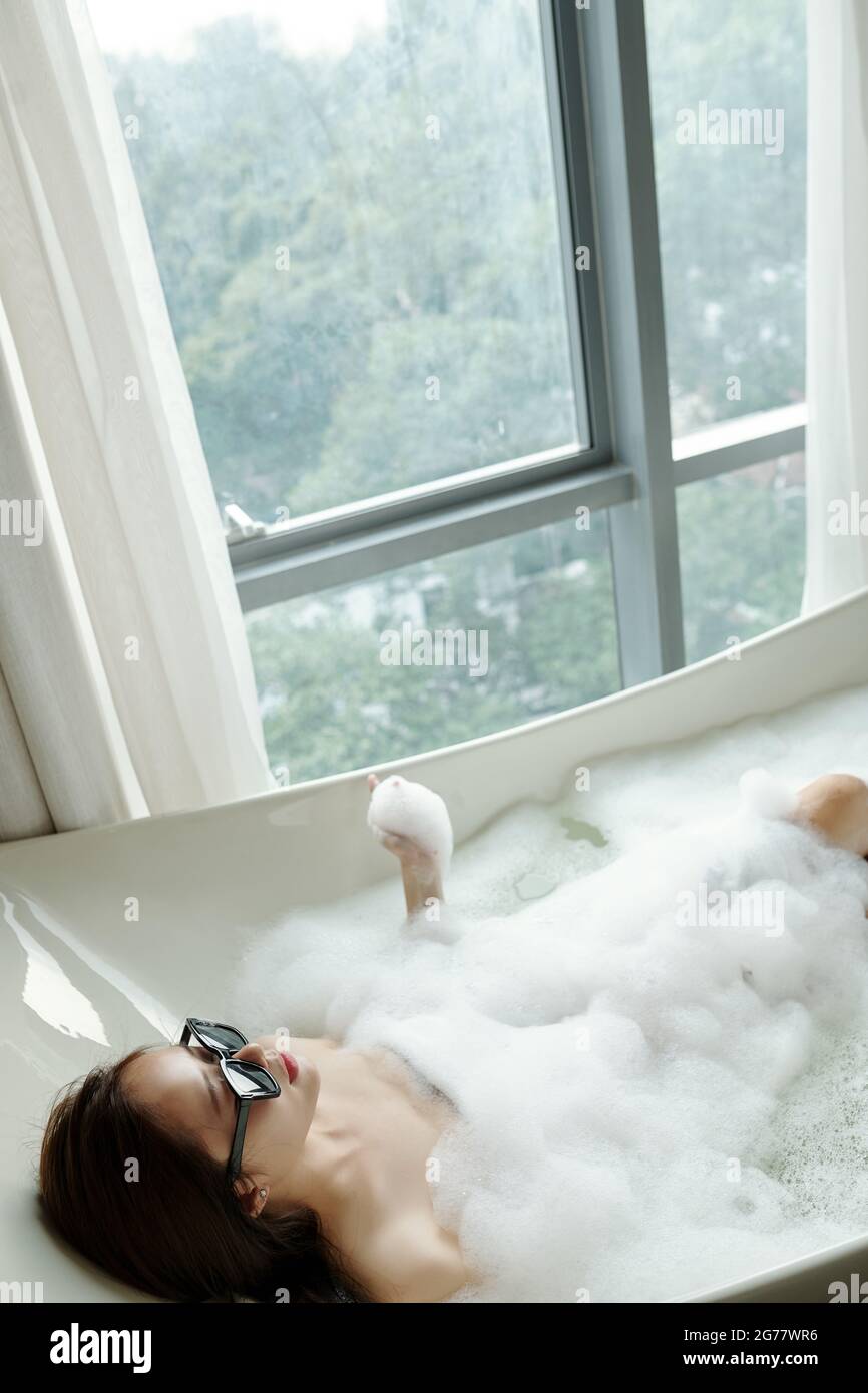 Junge Frau in Sonnenbrillen, die sich nach einem schweren Tag im Bad mit Schaum entspannt Stockfoto