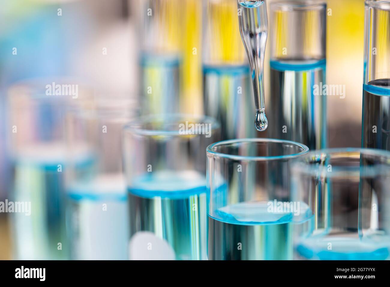Tropfen klarer blauer Flüssigkeit vom Tropfen zum Reagenzglas im Labor mit heller Farbunschärfe im Hintergrund. Stockfoto