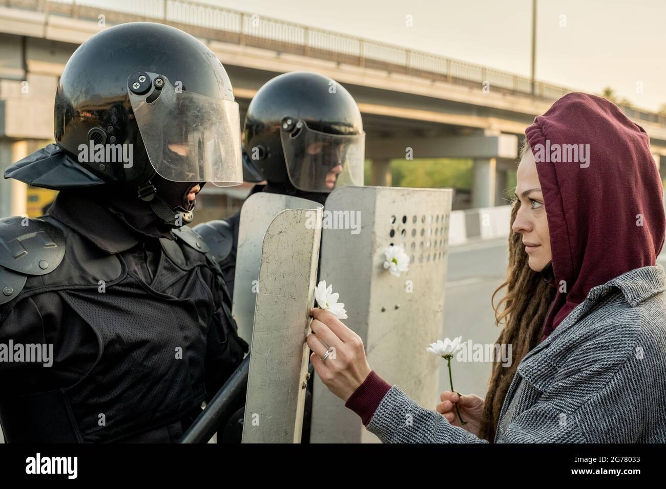 Friedliebende junge Frau in der Kapuze, die vor der Polizei steht, die bei der Rebellion Gänseblümchen an Schilde befestigt Stockfoto