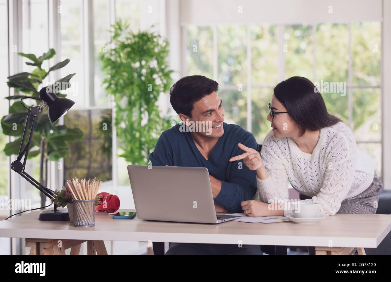 Der kaukasische Mann und die asiatische Frau mischen sich zwischen Rennliebhabern, die über einen Laptop-Notebook-Computer zusammenarbeiten und im Wohnzimmer mit glücklichen und intimen Menschen diskutieren. Neu Stockfoto